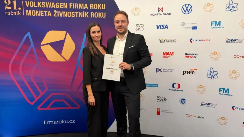 Zwei Personen mit der LOMAX-Urkunde vor der Veranstaltungswand Firma roku 2026